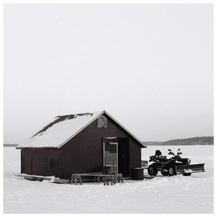 A small snow-covered hut with an open door stands next to an ATV with a snowplow on a vast expanse of snow.