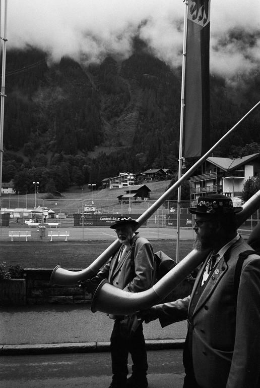 Two men in traditional attire carry long horns on their shoulders, with a mountain and village in the background.
