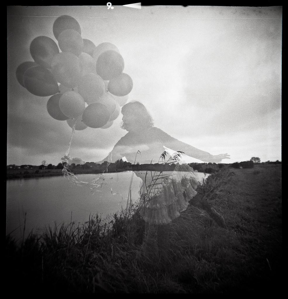 A double exposure shows a woman holding balloons superimposed over a landscape with a body of water and tall grass.