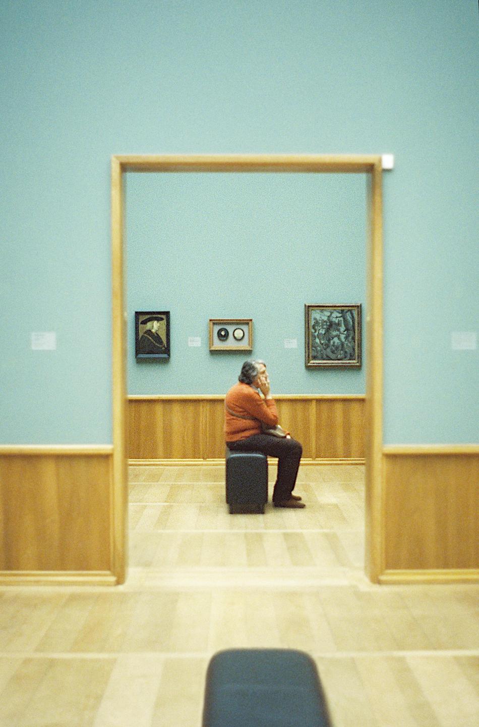 A man sits on a stool in a museum gallery with paintings on the wall.