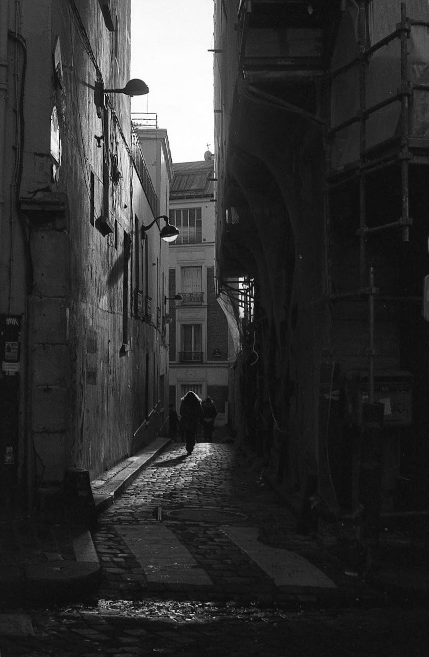 A person walks down a narrow, cobbled alleyway between tall buildings, with sunlight illuminating the path ahead.