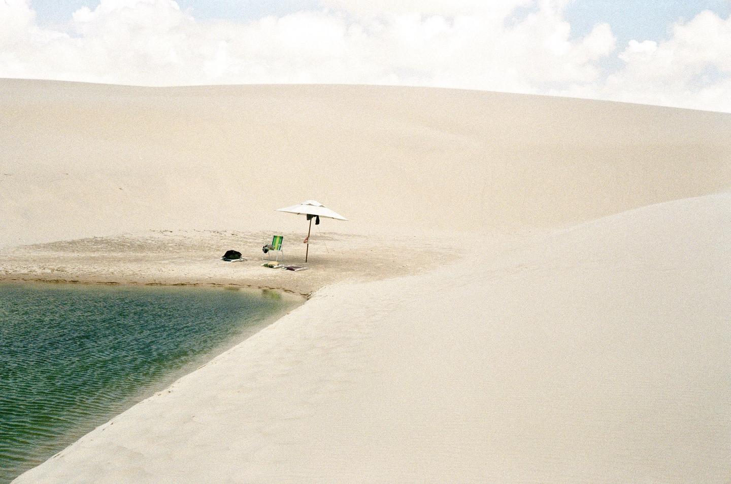 A white umbrella and a green chair sit on a sandy bank next to a body of water, surrounded by sand dunes under a cloudy sky.