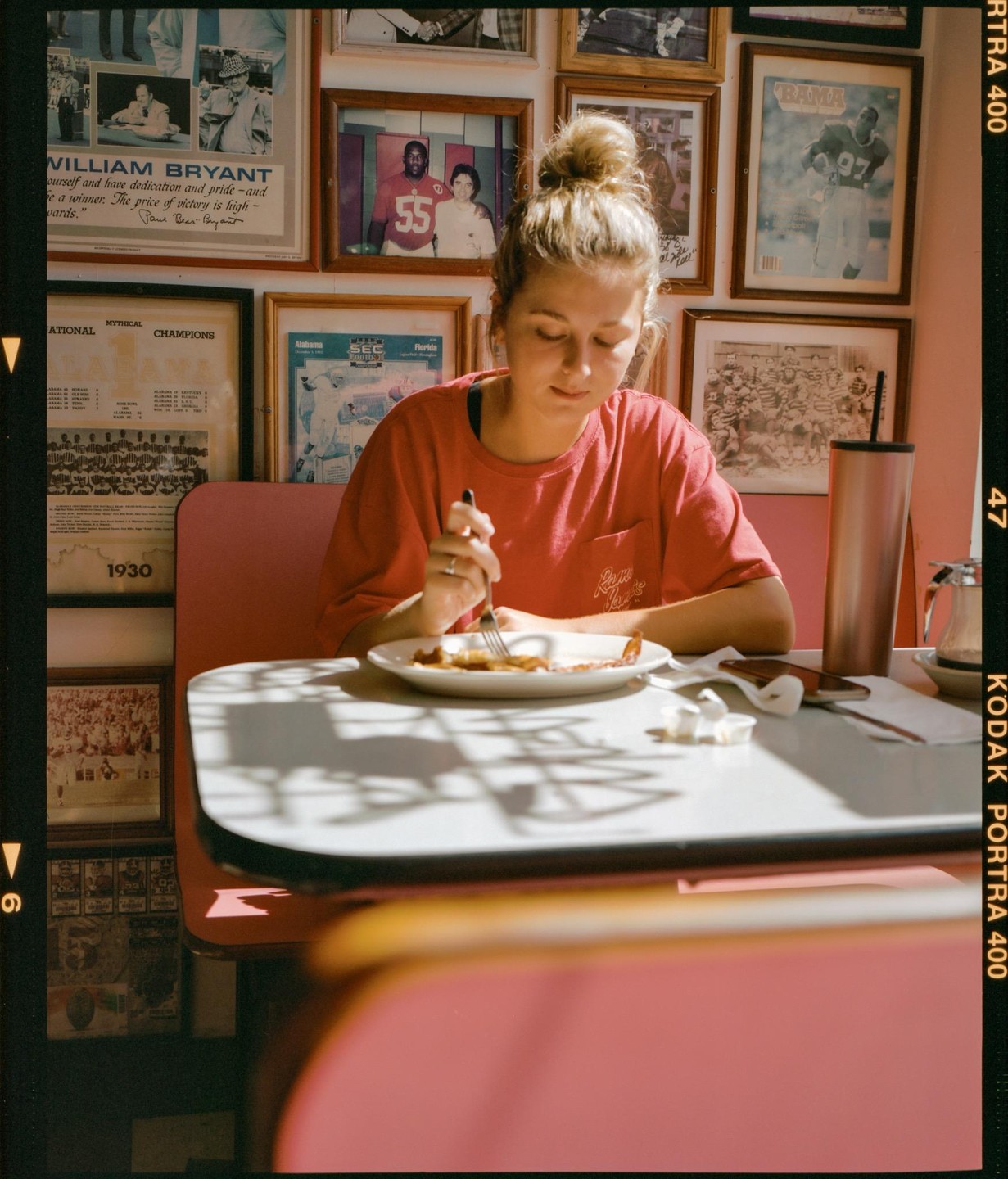 A woman with her hair in a bun eats breakfast at a diner booth with framed pictures on the wall.