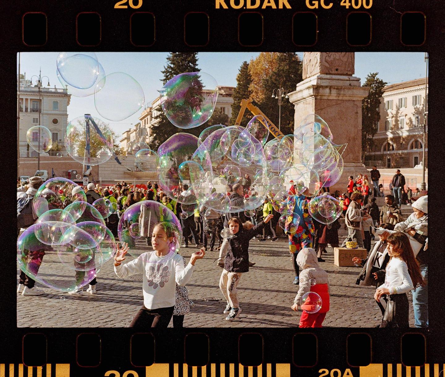 Children play with bubbles in a crowded city square with historic buildings and a monument in the background.