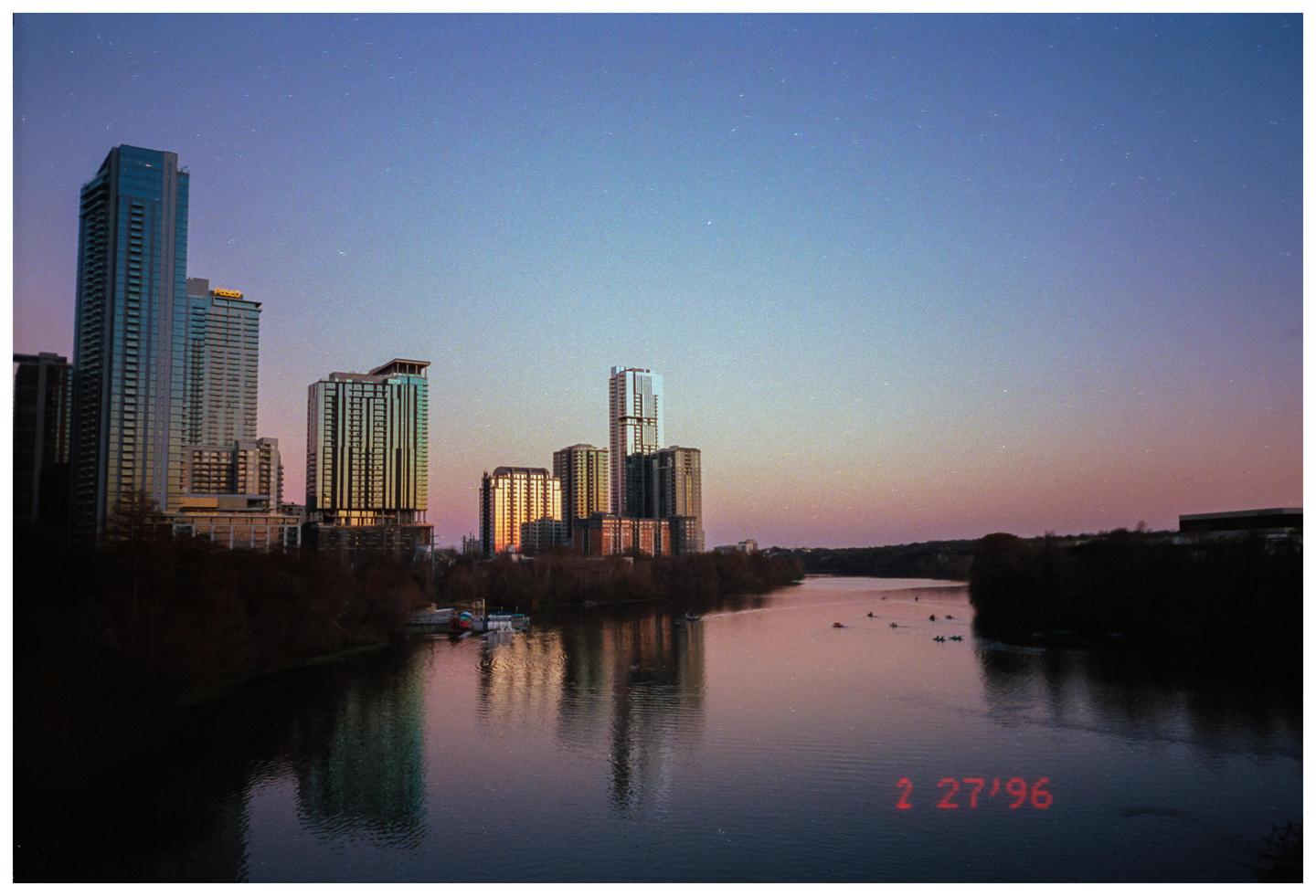 A cityscape with tall buildings is reflected in a river at dusk.
