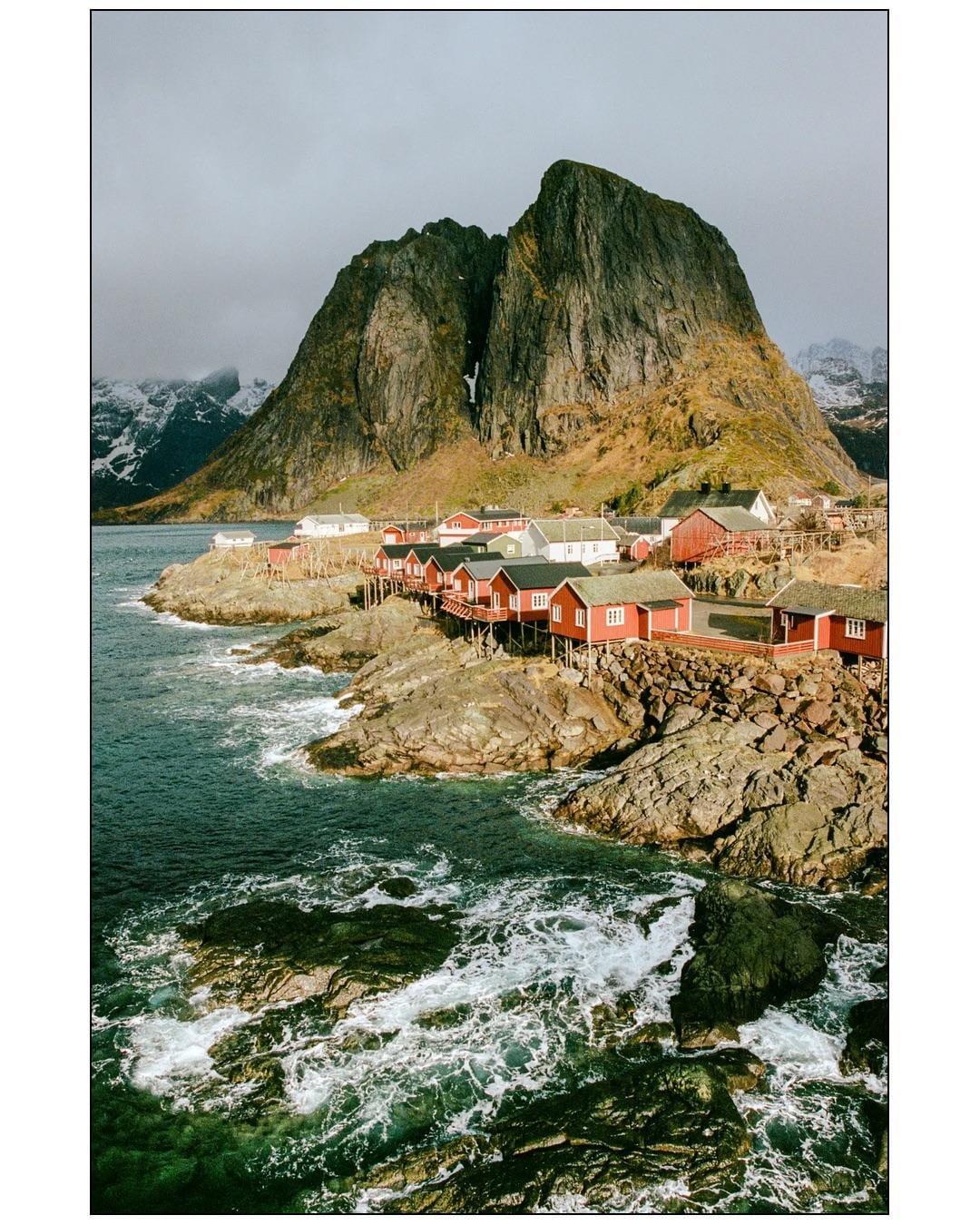 Red and white houses on stilts line a rocky coast with a large mountain in the background.
