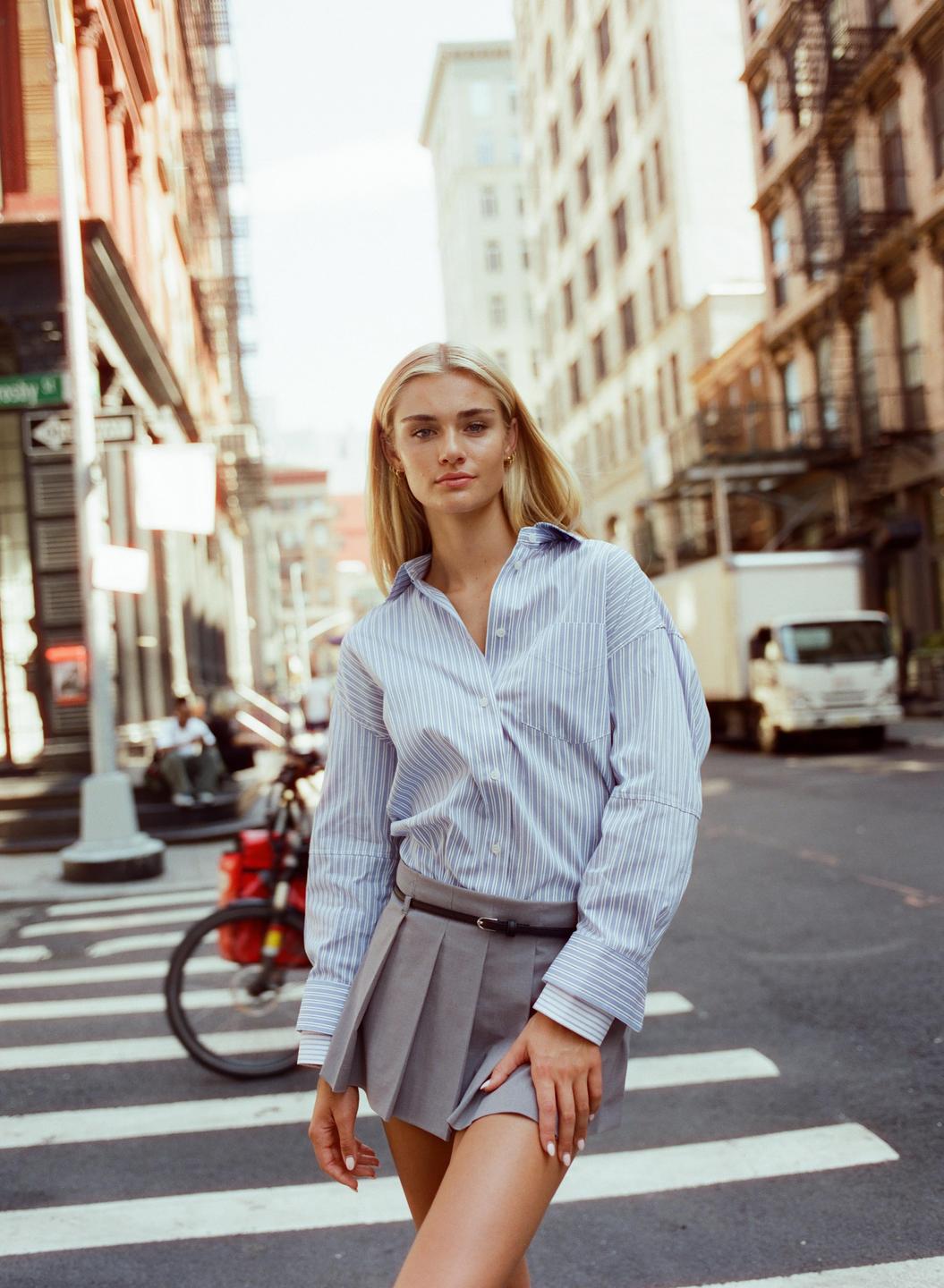A woman wearing a striped shirt and pleated skirt stands in the middle of a city street.