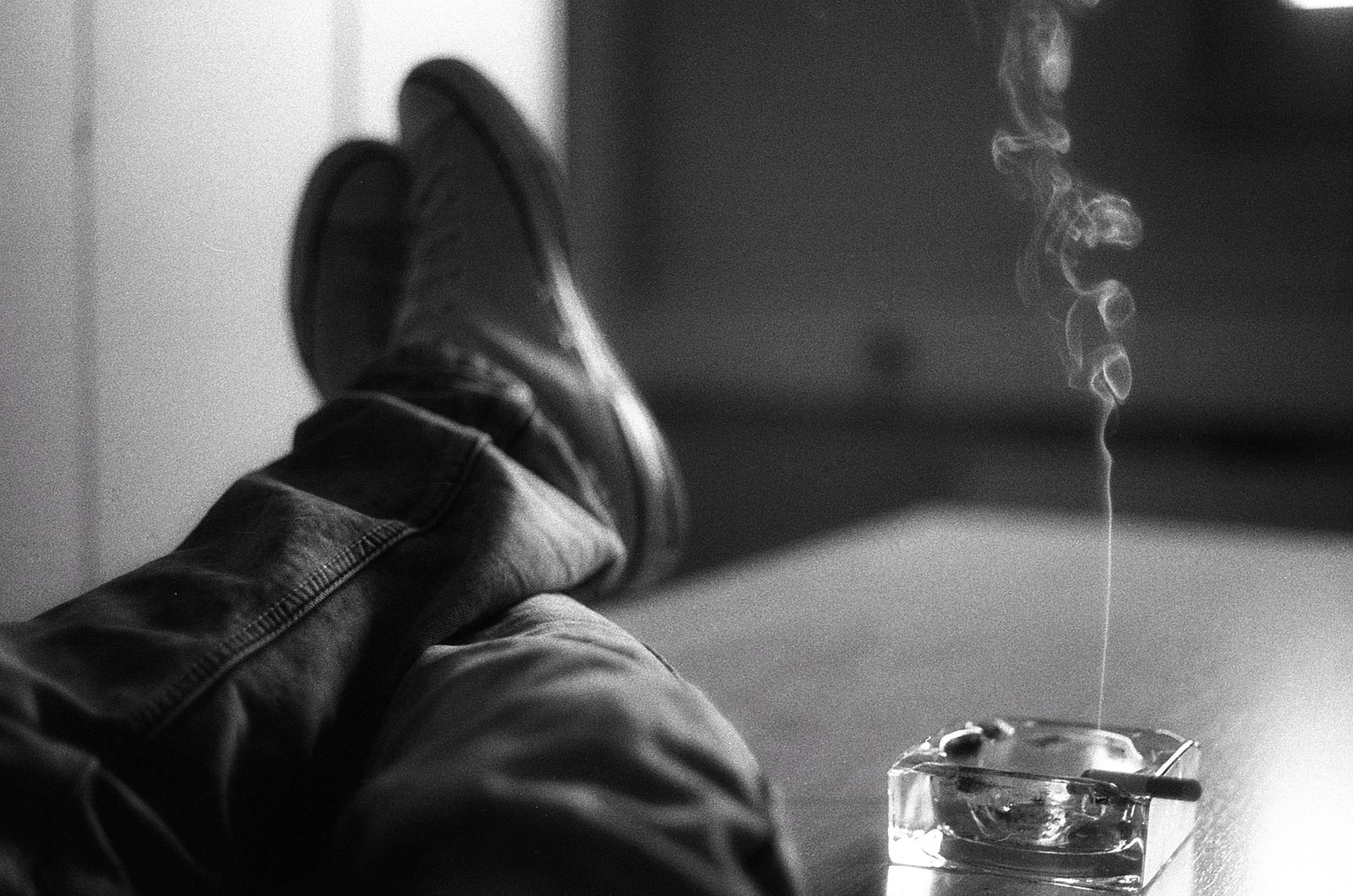 A person's legs are propped up next to an ashtray with a lit cigarette and rising smoke.