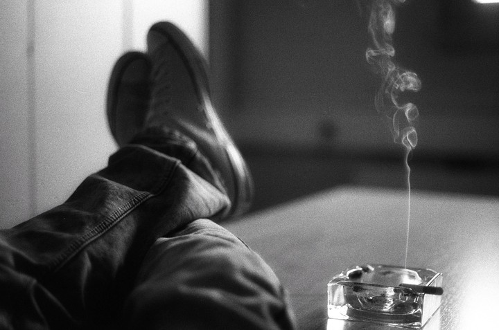 A person's legs are propped up next to an ashtray with a lit cigarette and rising smoke.
