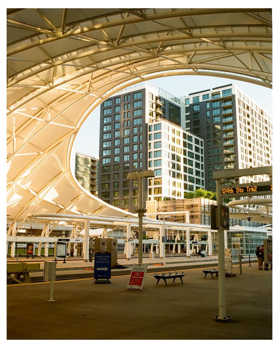 A train station platform is visible under a large arched canopy with tall buildings in the background.