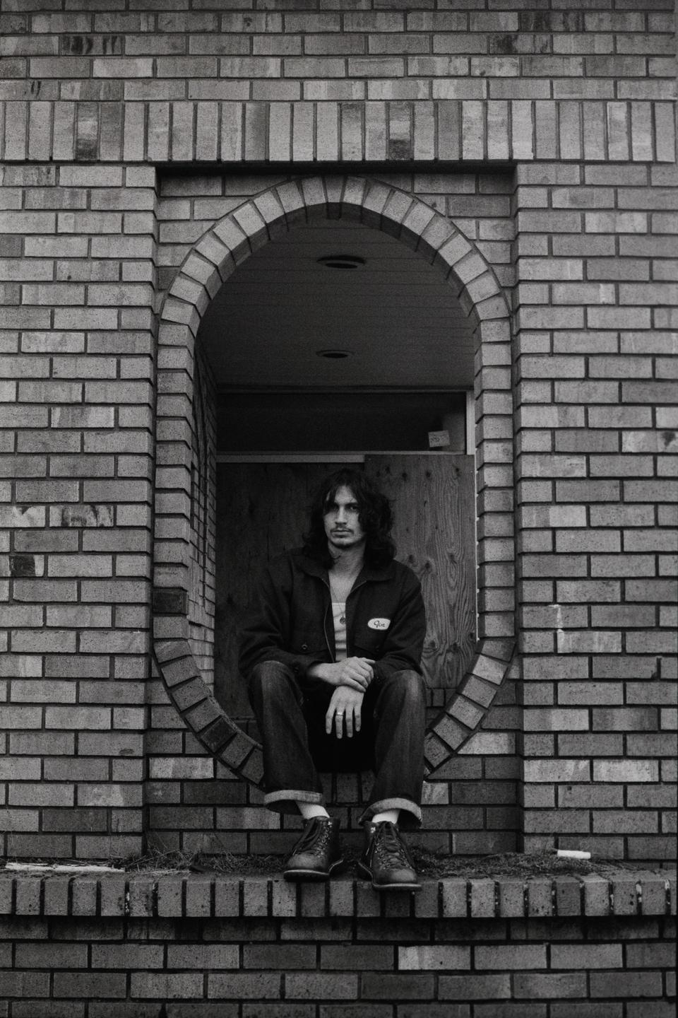 A man with long hair sits in an arched brick doorway.