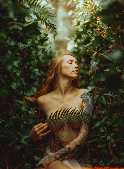 A woman with long hair and tattoos is partially covered by a fern leaf in a lush green setting.