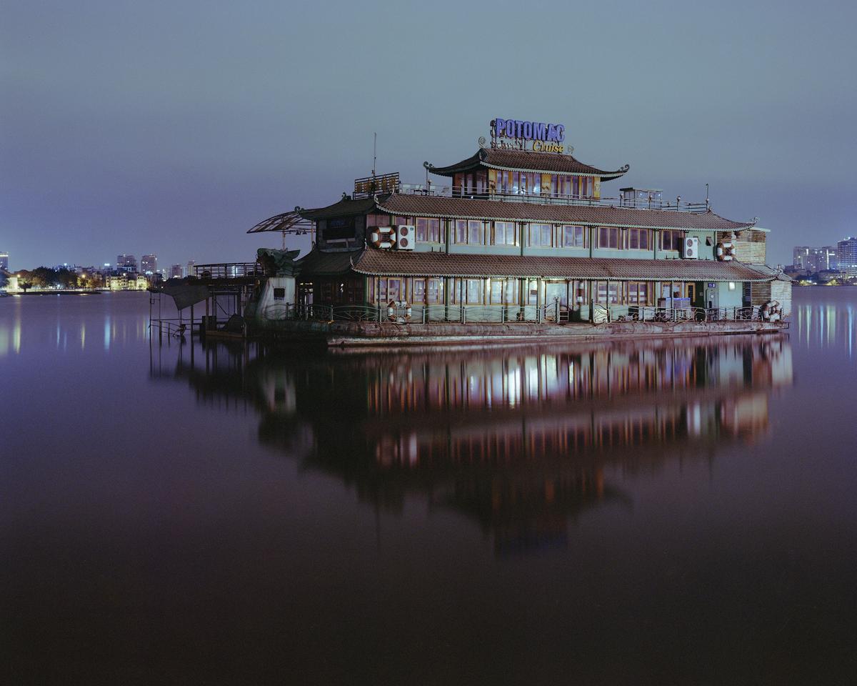 A large boat with a pagoda-style roof is illuminated at night, reflecting on the still water.