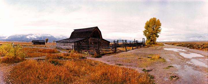A panoramic landscape shows a wooden barn and fence in a field with mountains in the background under a cloudy sky.