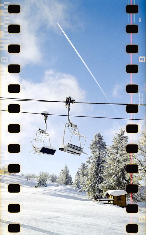Two empty ski lift chairs hang over a snow-covered mountain with pine trees and a small cabin under a blue sky with a contrail.