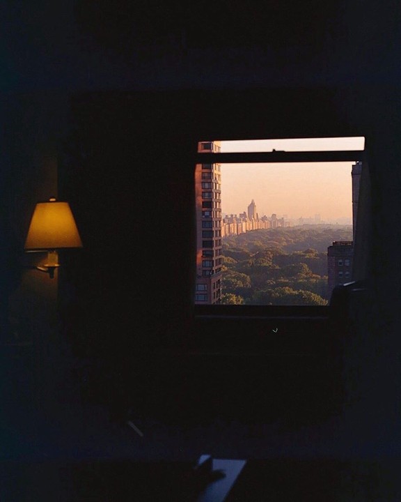 A window looks out onto Central Park and the New York City skyline at sunset, with a lamp visible in the dark room.