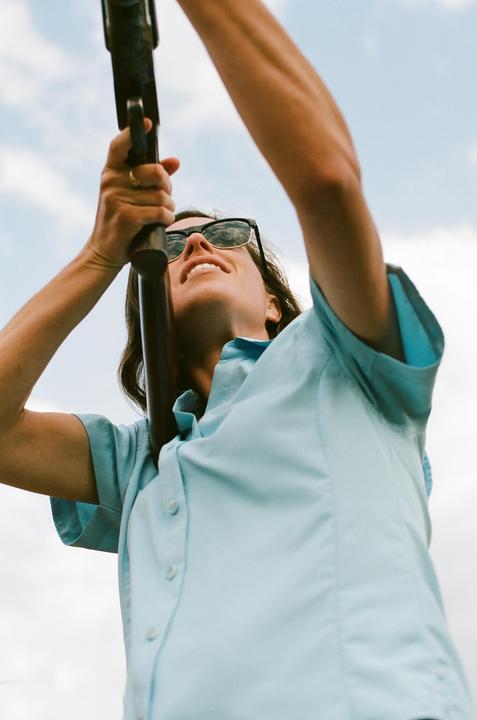 A woman wearing sunglasses and a light blue shirt holds a rifle pointed upwards against a cloudy sky.