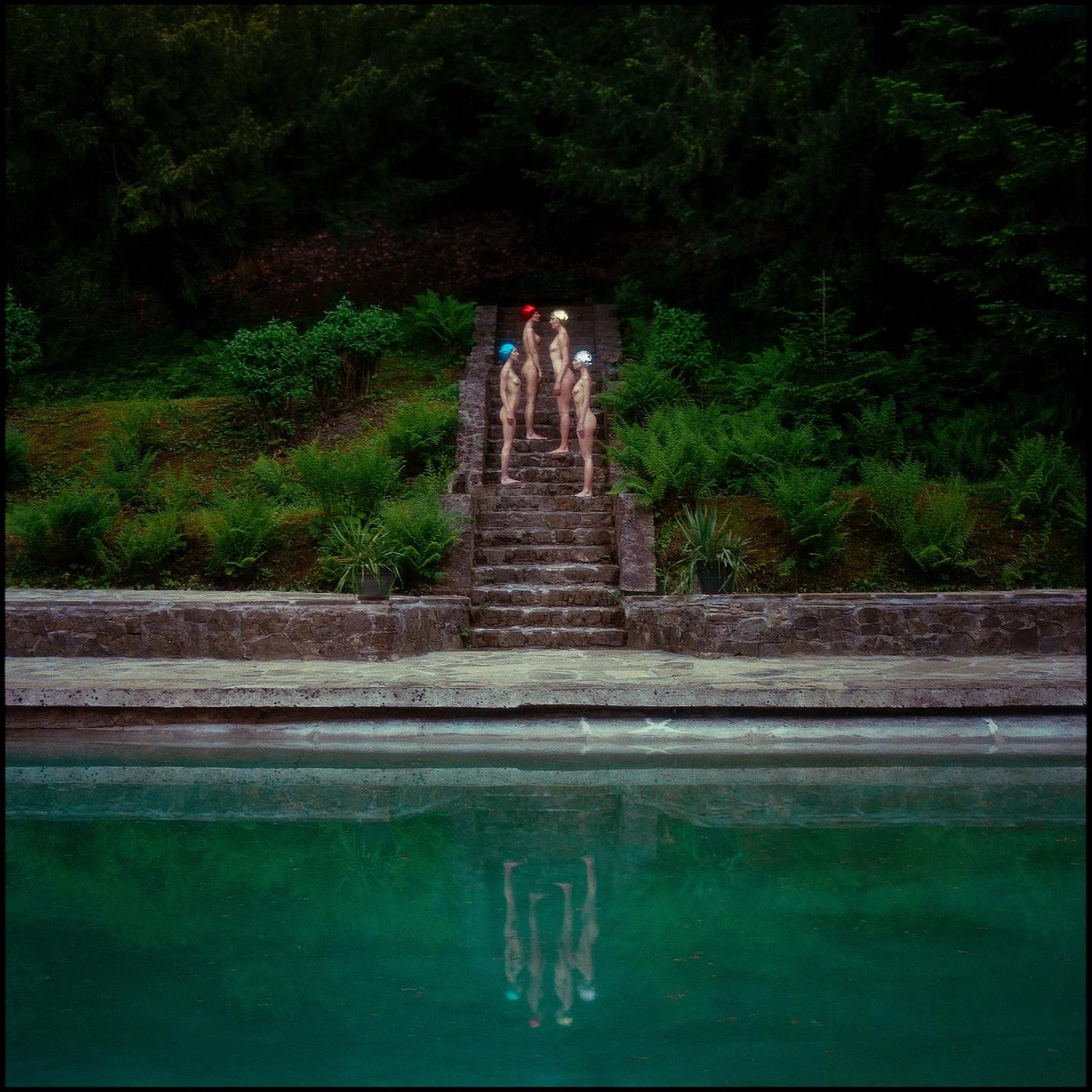 Four people wearing swimming caps stand on stone steps above a green swimming pool, with their reflections visible in the water.