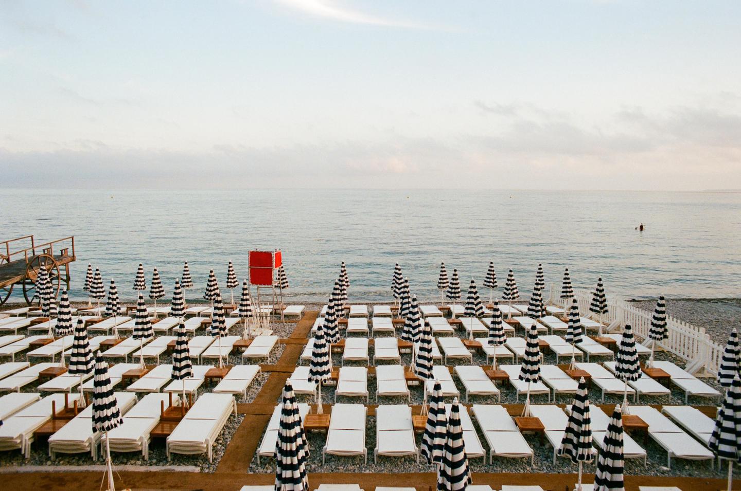 A beach is filled with rows of lounge chairs and striped umbrellas facing the ocean, with a red lifeguard stand in the middle.