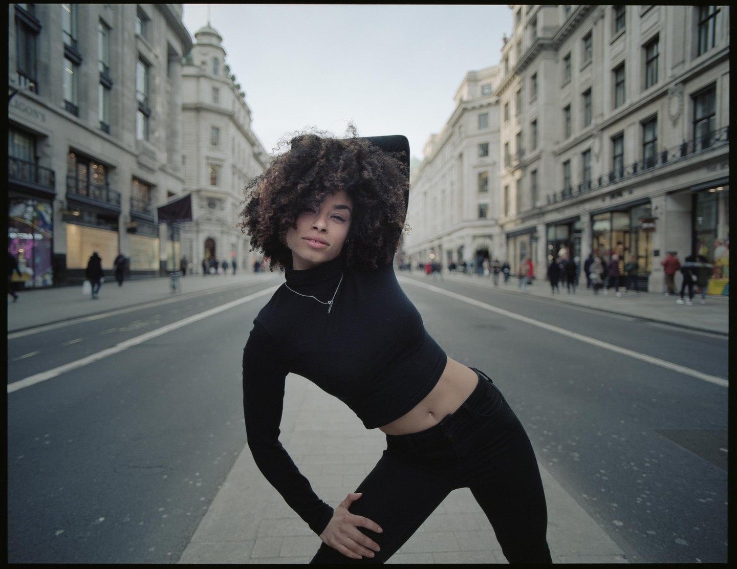 A woman with curly hair poses in the middle of a street lined with buildings.