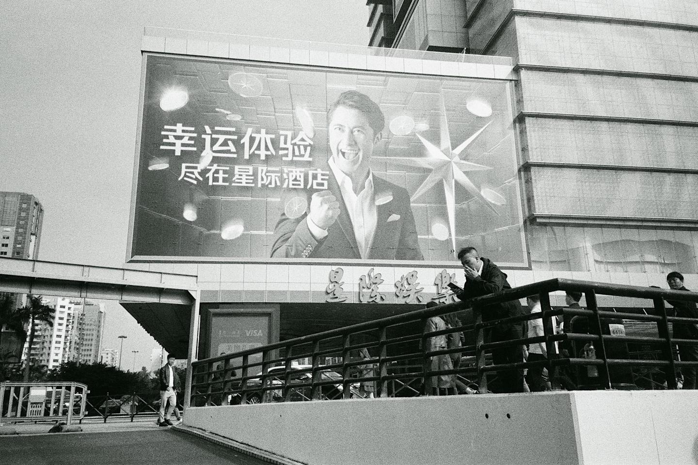A large billboard with a man in a suit smiling and clenching his fist is above a street with people and buildings.