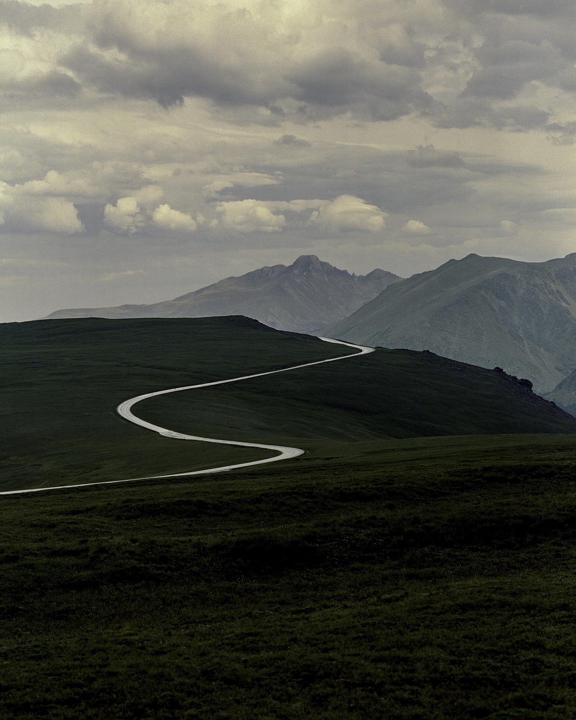 A winding road cuts through a green, hilly landscape under a cloudy sky, with mountains in the distance.