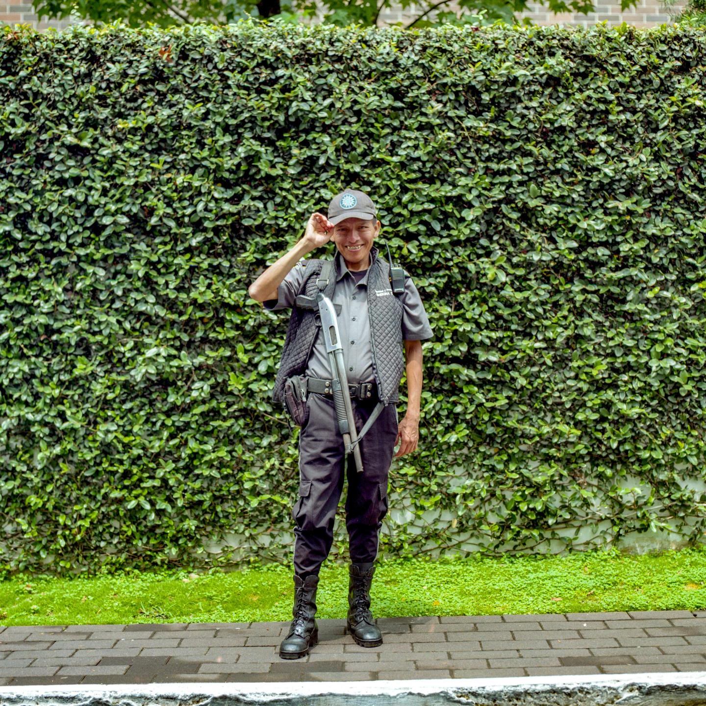 A man in a security uniform with a shotgun stands on a sidewalk in front of a lush green ivy wall.