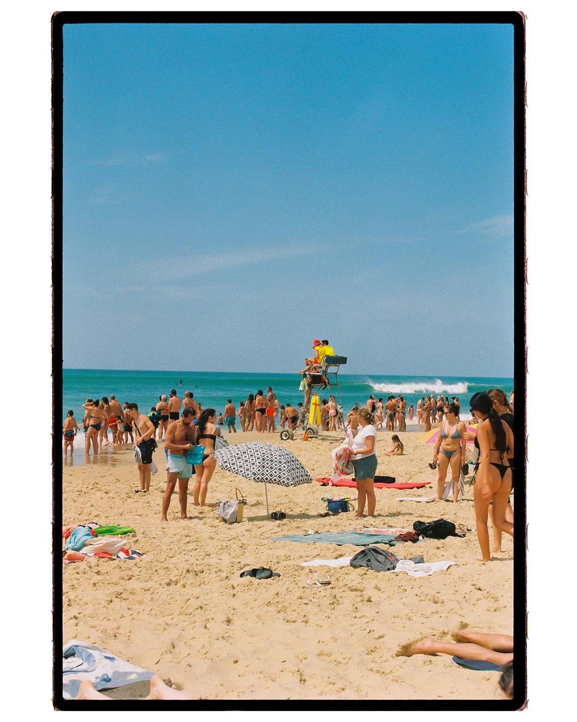 A crowded beach is visible with many people swimming in the ocean and sunbathing on the sand.
