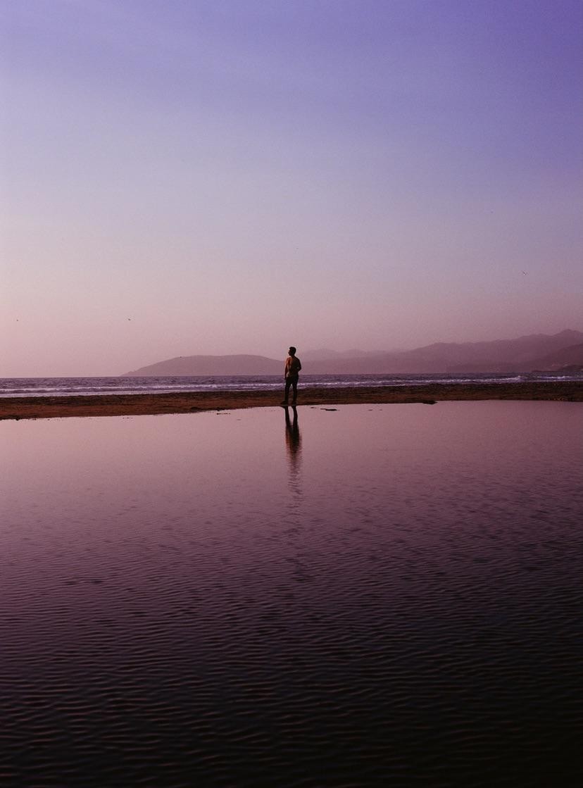 A man stands on a strip of land between a lagoon and the ocean, with mountains in the distance under a purple sky.