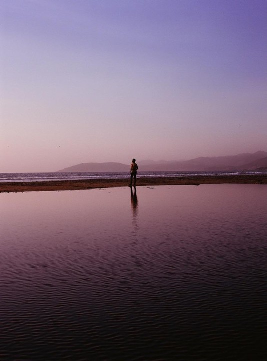 A man stands on a strip of land between a lagoon and the ocean, with mountains in the distance under a purple sky.
