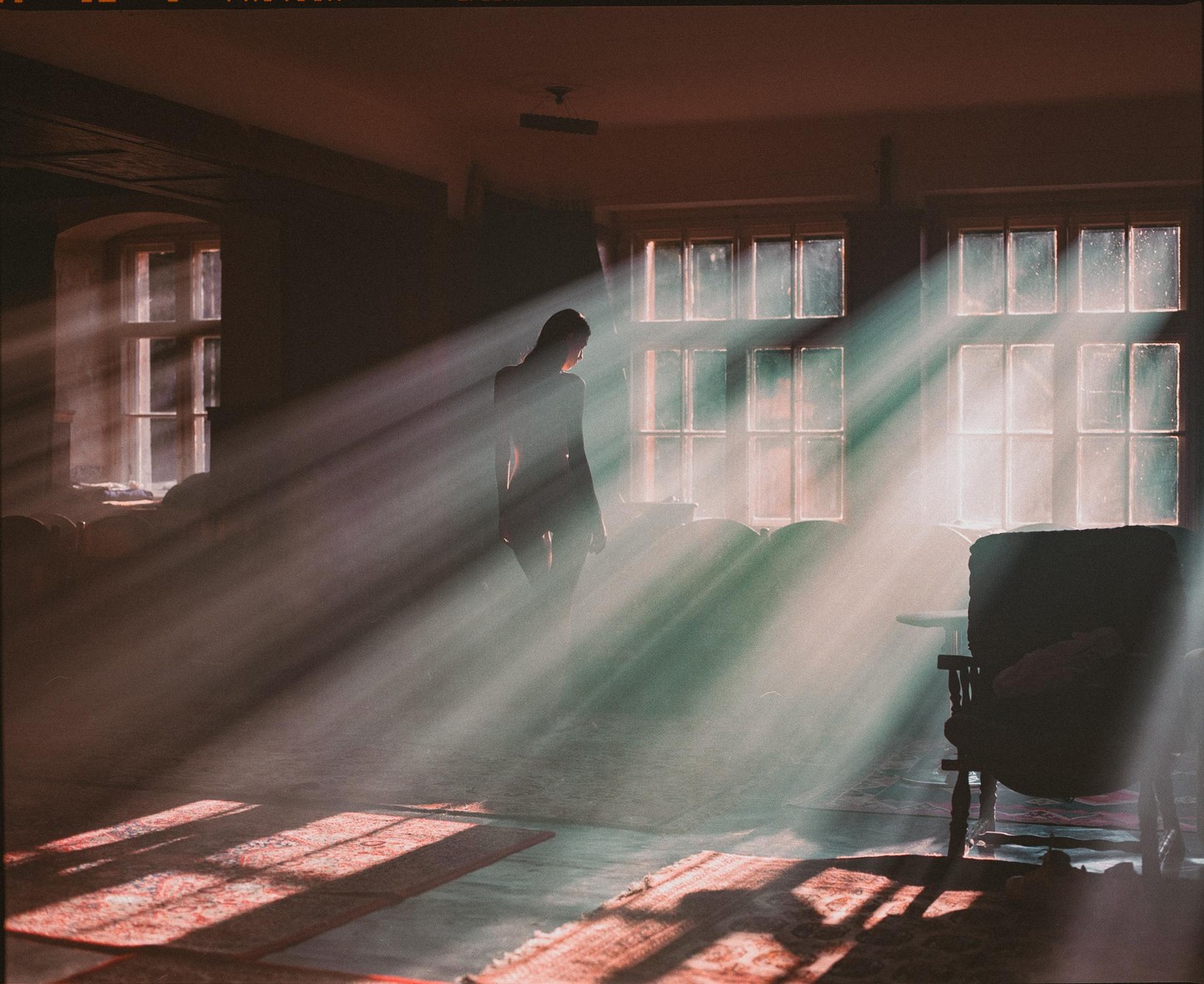 A woman stands in a sunlit room with light rays streaming through windows.