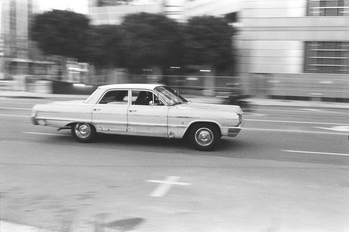 A white vintage car drives on a street with blurred buildings and trees in the background.