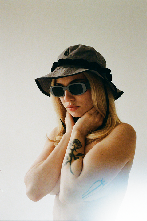 A woman wearing a bucket hat and sunglasses poses with her hands near her neck, revealing a rose tattoo on her arm.