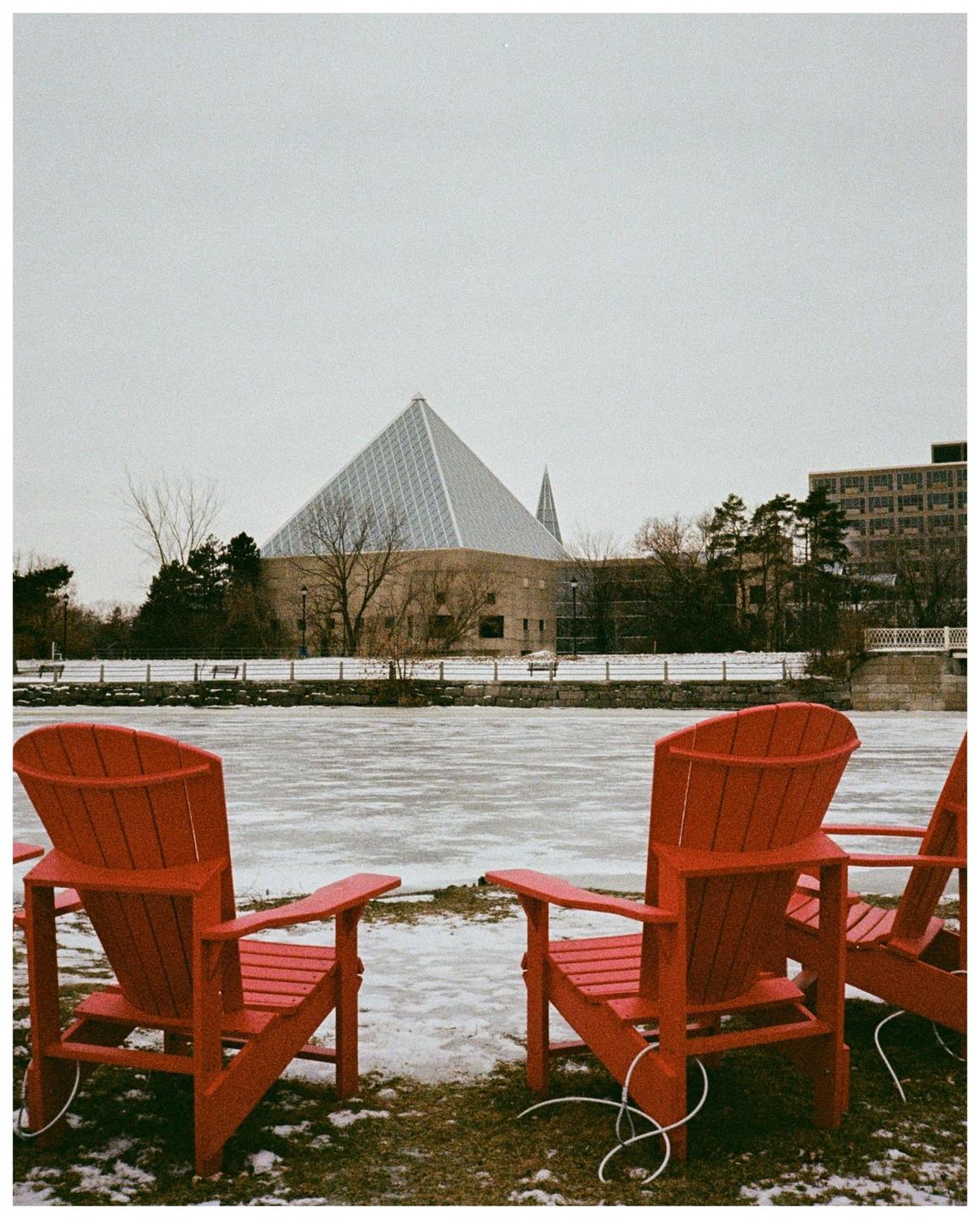 Three red Adirondack chairs sit on a snowy bank overlooking a frozen canal with a pyramid-shaped building in the background.