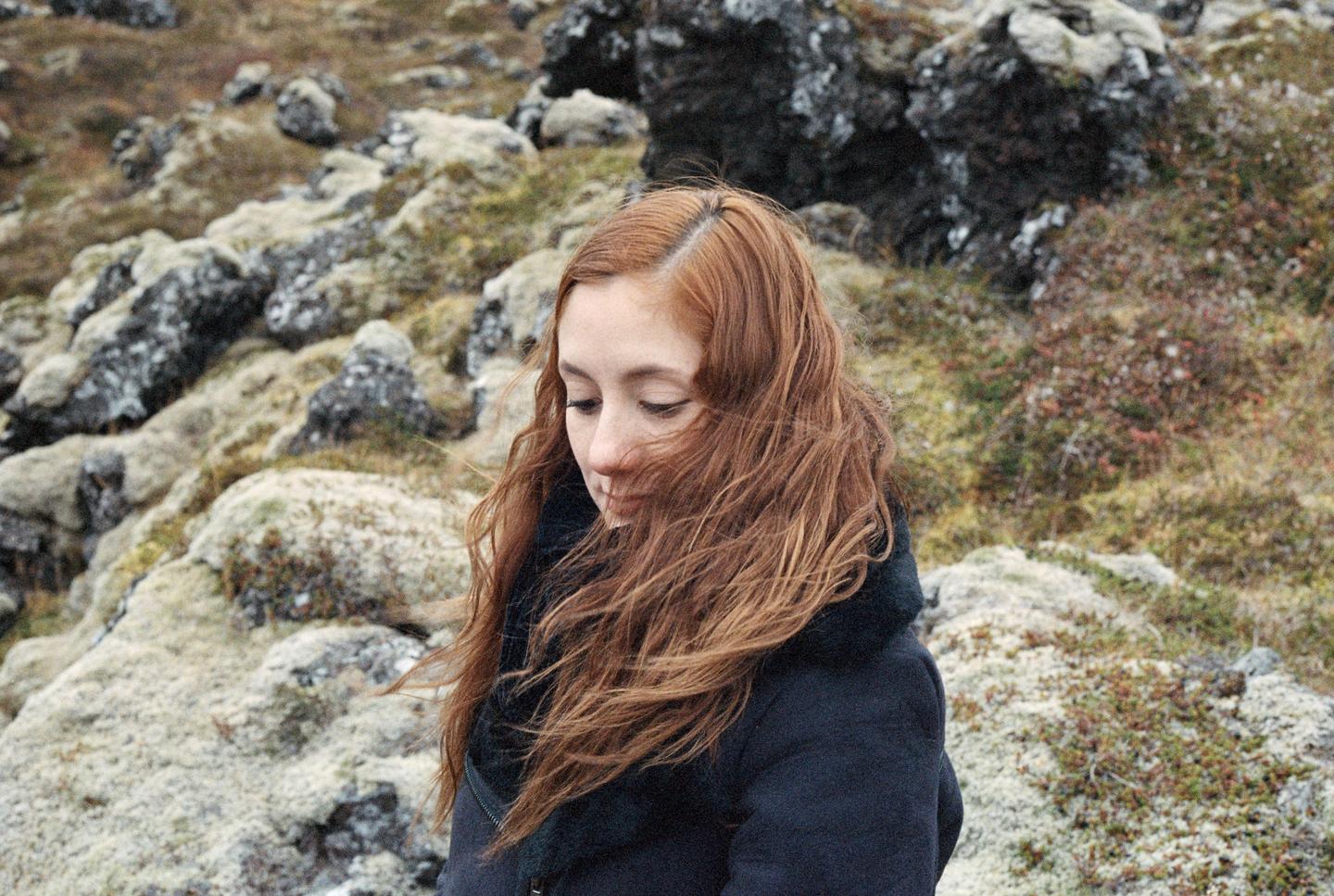 A woman with long, wavy red hair looks down while standing in a rocky, moss-covered landscape.