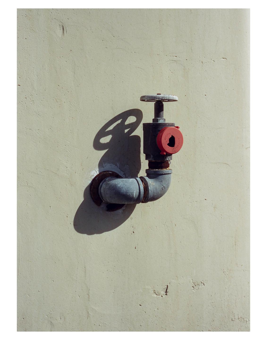 A pipe with a red cap and a valve wheel is attached to a textured wall, casting a shadow.