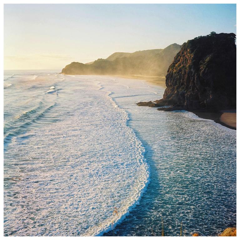 Waves crash on a beach next to a large rock formation and hills in the distance.