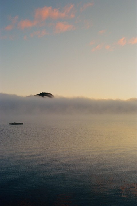 A misty lake with a small floating dock and a distant hill under a sky with pink clouds.