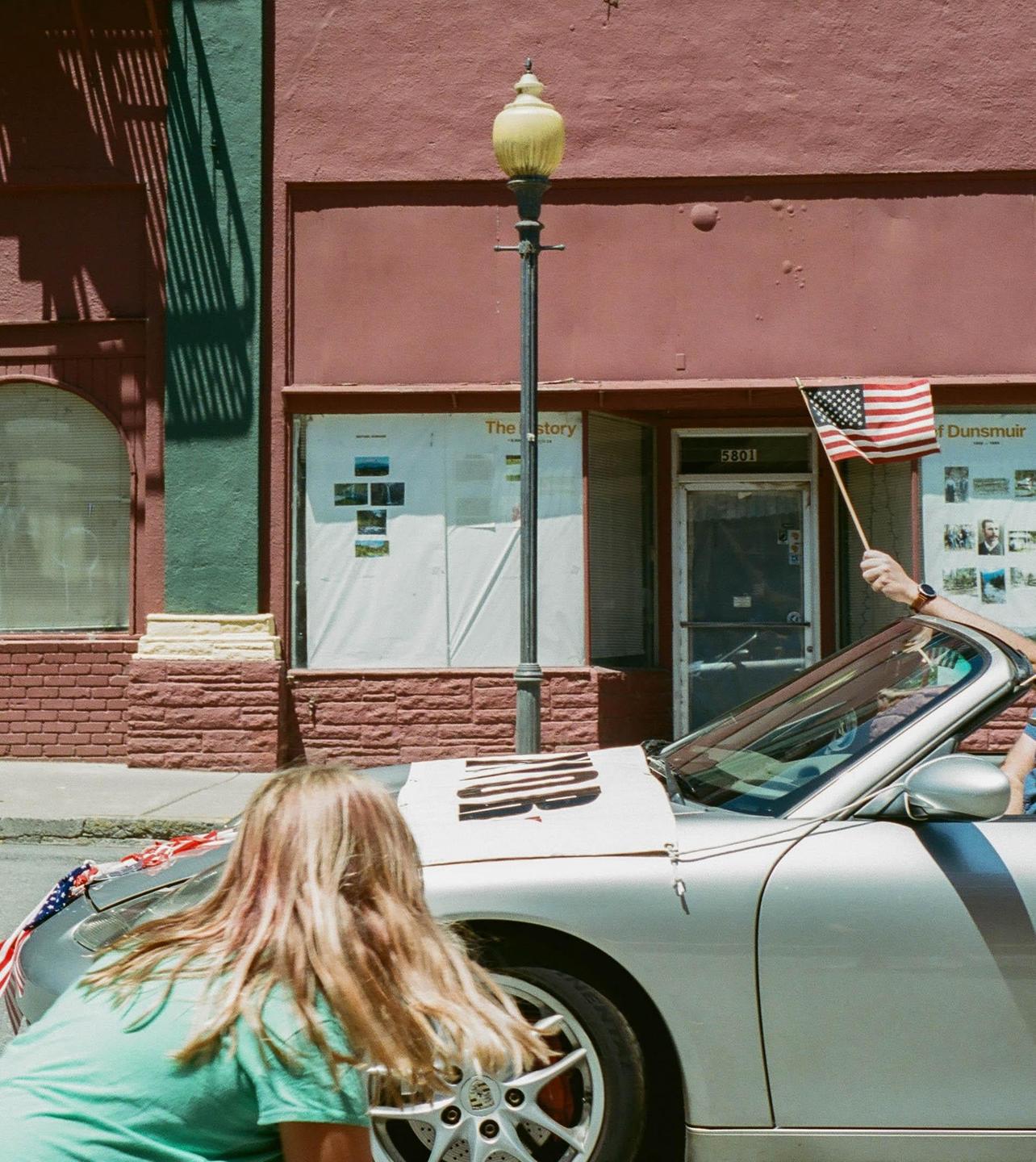 A woman bends over the front of a silver convertible car while a hand holds a small American flag out of the passenger side window.