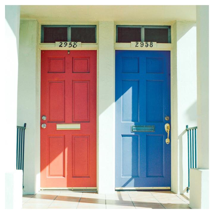 Two doors, one red and one blue, are side by side with sunlight and shadow falling across them.