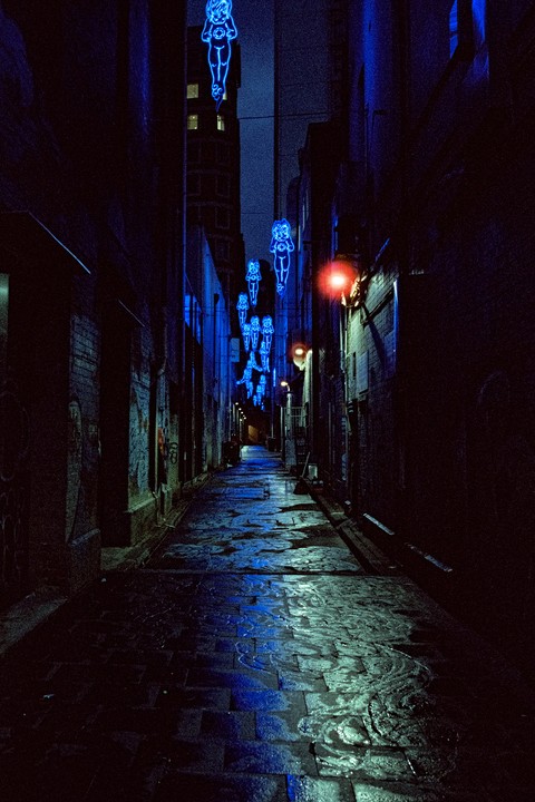A narrow alleyway at night is illuminated by blue neon figures hanging overhead and reflected on the wet pavement.