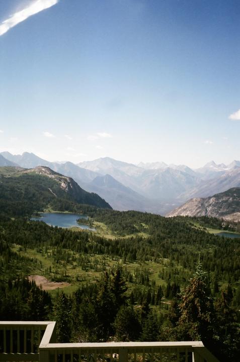 A vast mountain landscape features a valley filled with evergreen trees and two lakes, all under a clear sky.