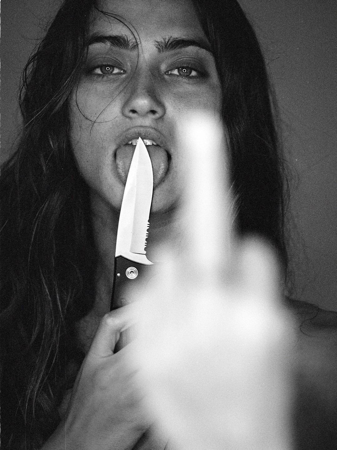 A woman holds a knife to her tongue while a blurred hand is in the foreground.