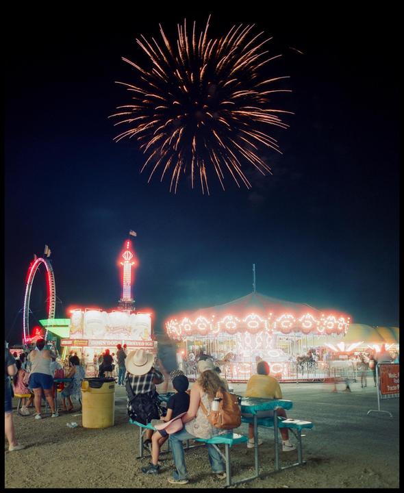 A firework explodes over a carnival with people sitting on benches in the foreground.
