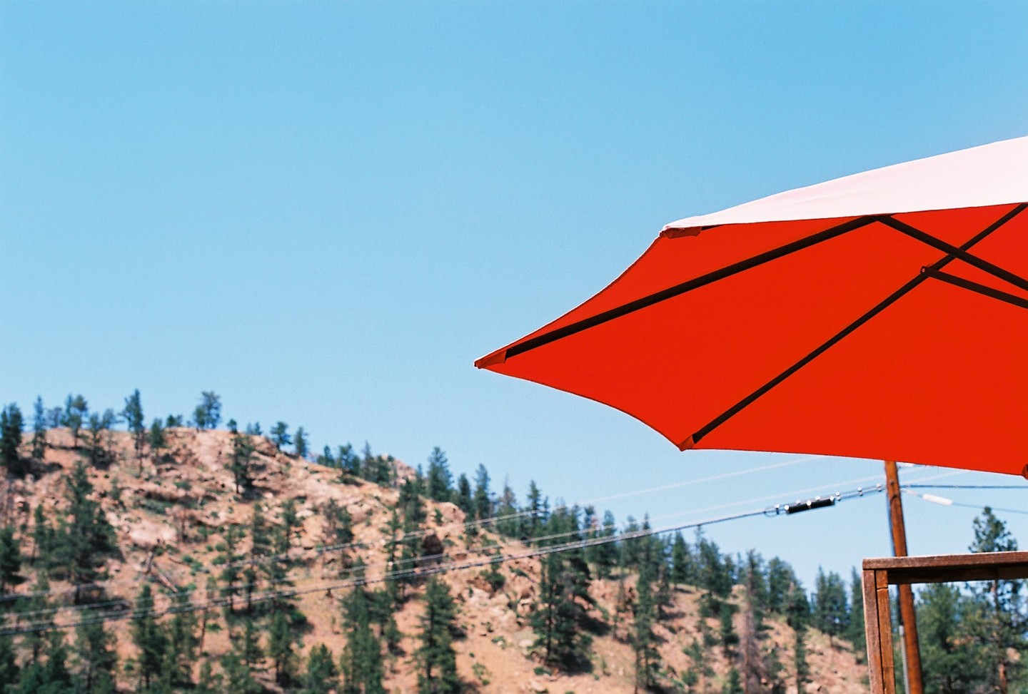 A bright red and white umbrella is in the foreground with a tree-covered hill under a clear blue sky in the background.