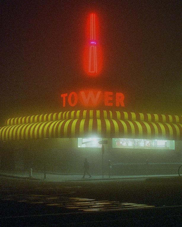 A person walks past a building with a striped awning and a neon sign that reads "TOWER" in the fog at night.
