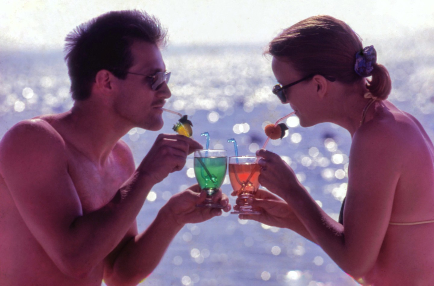 A man and a woman drink colorful cocktails with straws by the water.