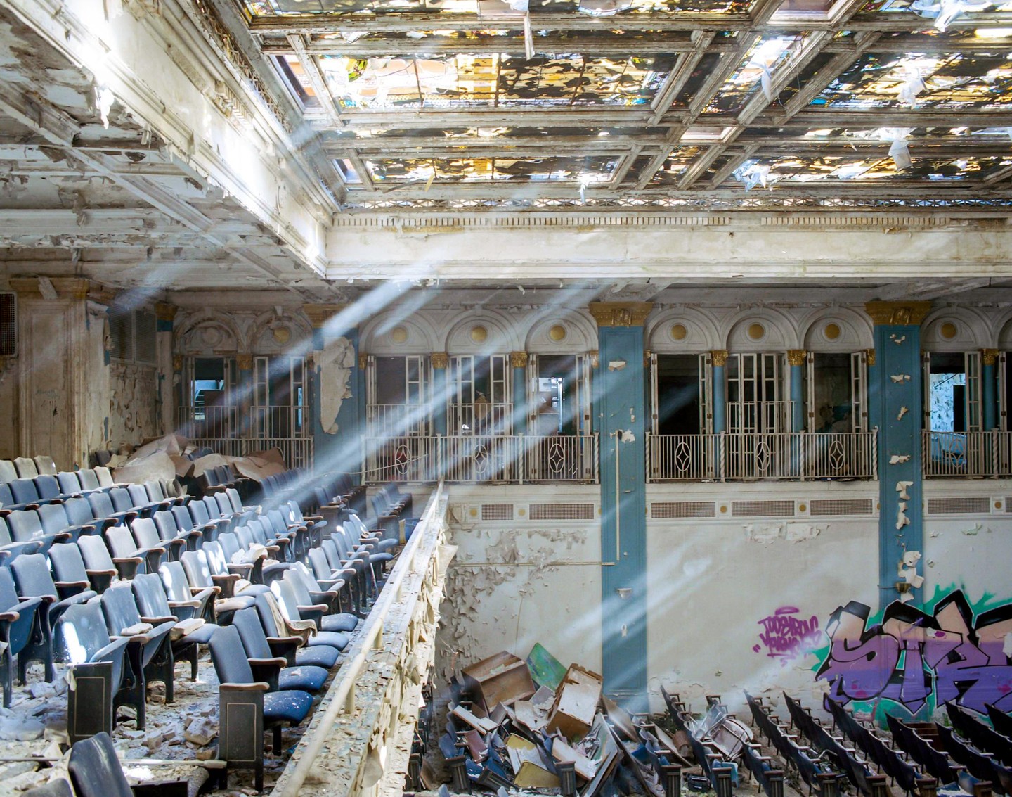Light rays shine through the stained glass ceiling of an abandoned theater with rows of seats and graffiti on the wall.