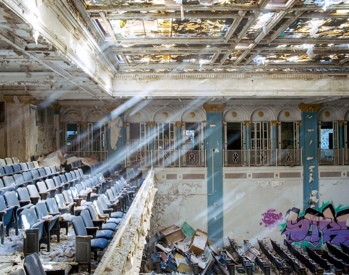 Light rays shine through the stained glass ceiling of an abandoned theater with rows of seats and graffiti on the wall.