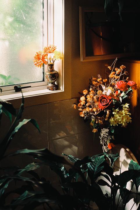 A vase of orange flowers sits on a window sill next to a larger bouquet of red, yellow, and orange flowers, with green foliage in the foreground.